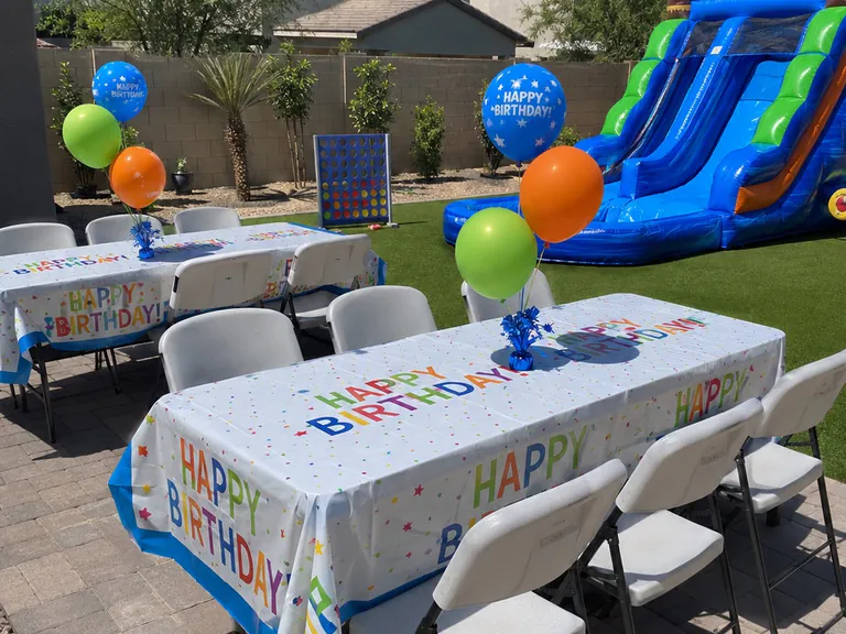 Backyard birthday party setup with tables, white folding chairs, balloons, giant Connect 4, and an inflatable slide